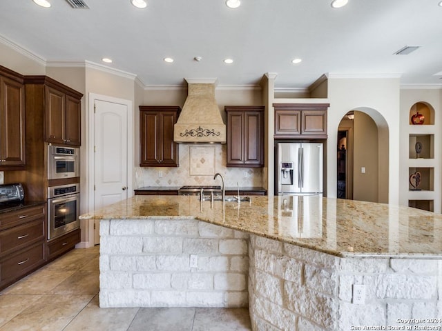 kitchen with appliances with stainless steel finishes, light stone countertops, custom exhaust hood, a large island with sink, and a sink