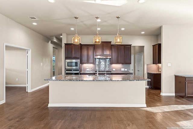 kitchen featuring pendant lighting, dark wood-type flooring, an island with sink, appliances with stainless steel finishes, and tasteful backsplash