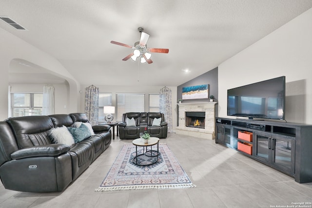 tiled living room with ceiling fan, a stone fireplace, a textured ceiling, and vaulted ceiling