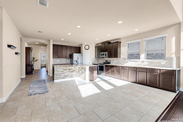 kitchen featuring light tile patterned floors, an island with sink, tasteful backsplash, dark brown cabinets, and stainless steel appliances