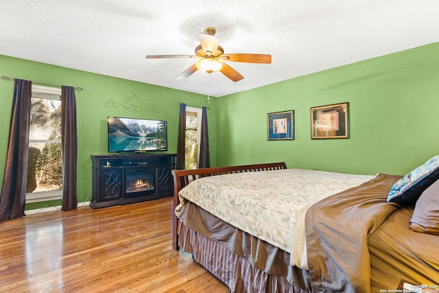 bedroom with a glass covered fireplace, ceiling fan, light wood-style flooring, and a textured ceiling