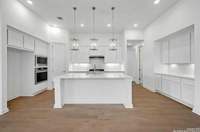 kitchen featuring decorative backsplash, appliances with stainless steel finishes, a kitchen island with sink, light hardwood / wood-style flooring, and hanging light fixtures