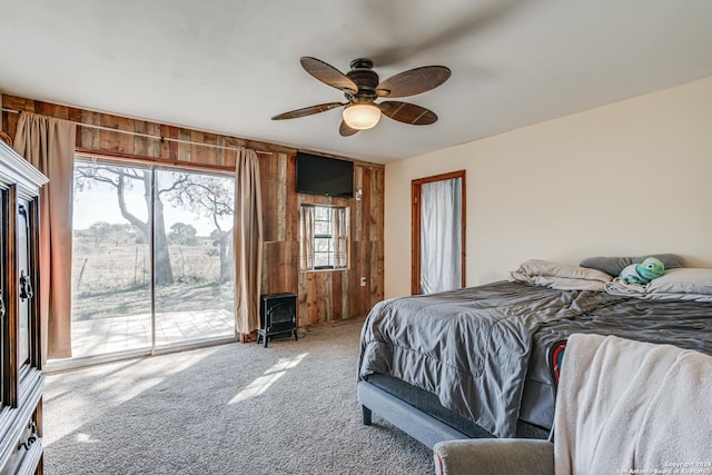 carpeted bedroom featuring access to outside, a wood stove, ceiling fan, and wood walls