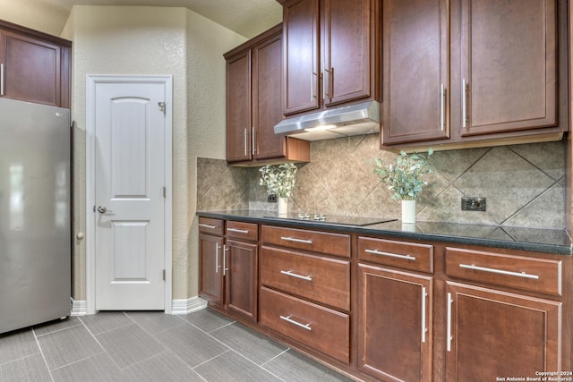 kitchen with black electric stovetop, decorative backsplash, dark stone countertops, and stainless steel refrigerator