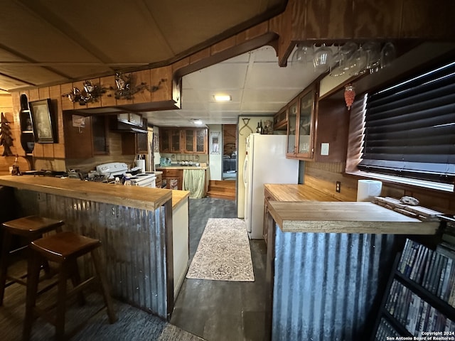 kitchen with a kitchen bar, wall chimney exhaust hood, white appliances, dark hardwood / wood-style floors, and wood walls