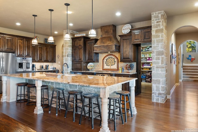 kitchen featuring hanging light fixtures, stainless steel appliances, tasteful backsplash, custom range hood, and a large island with sink