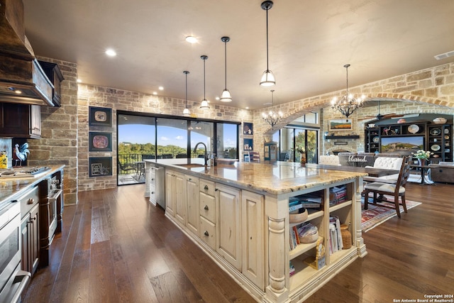 kitchen with a stone fireplace, an island with sink, sink, light stone counters, and dark wood-type flooring