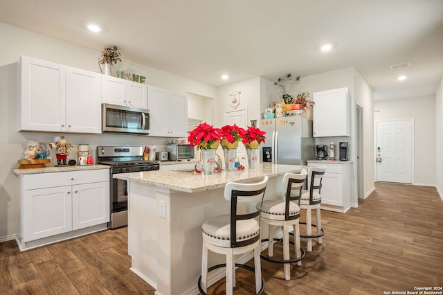 kitchen with appliances with stainless steel finishes, dark hardwood / wood-style flooring, light stone counters, white cabinets, and an island with sink