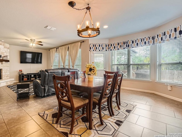 tiled dining space featuring ceiling fan with notable chandelier, a healthy amount of sunlight, and a stone fireplace