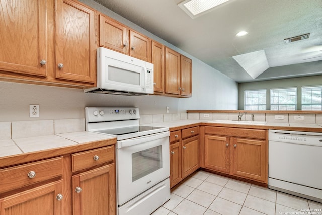 kitchen featuring vaulted ceiling with skylight, white appliances, sink, light tile patterned floors, and tile counters