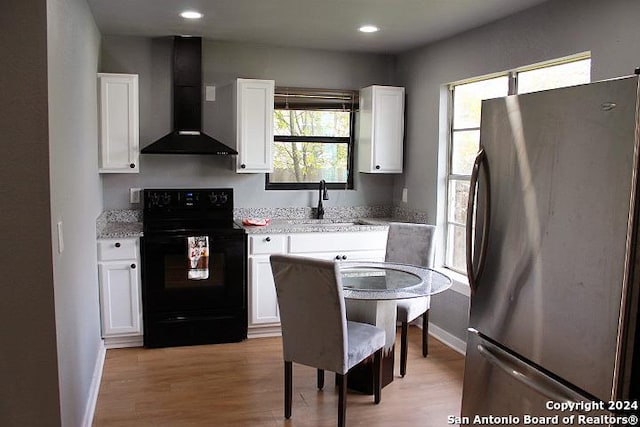 kitchen with stainless steel refrigerator, electric range, wall chimney exhaust hood, white cabinets, and light wood-type flooring