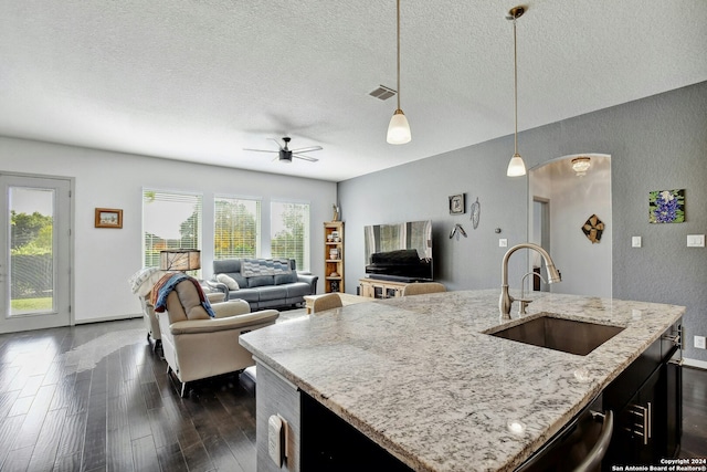 kitchen with dark wood-type flooring, a center island with sink, sink, hanging light fixtures, and light stone counters