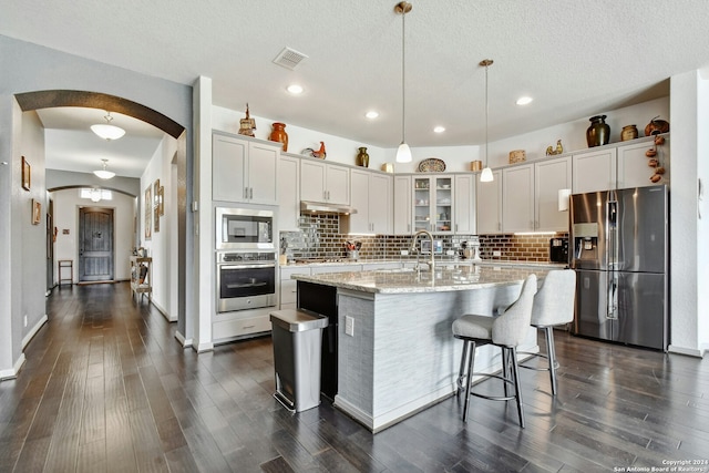 kitchen featuring dark wood-type flooring, hanging light fixtures, light stone counters, a kitchen island with sink, and appliances with stainless steel finishes