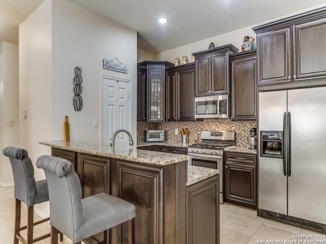 kitchen with dark brown cabinetry, stainless steel appliances, a kitchen breakfast bar, light stone counters, and light tile patterned flooring