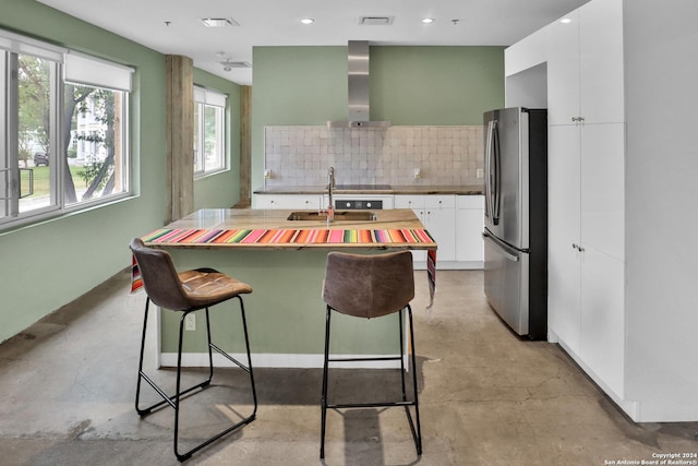 kitchen with a kitchen breakfast bar, backsplash, wall chimney exhaust hood, white cabinetry, and stainless steel refrigerator