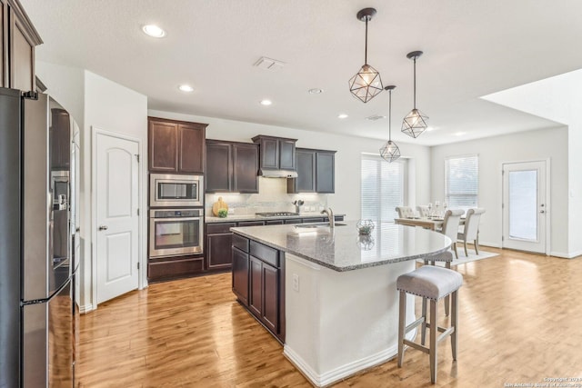 kitchen with dark brown cabinets, stainless steel appliances, decorative light fixtures, light hardwood / wood-style floors, and an island with sink