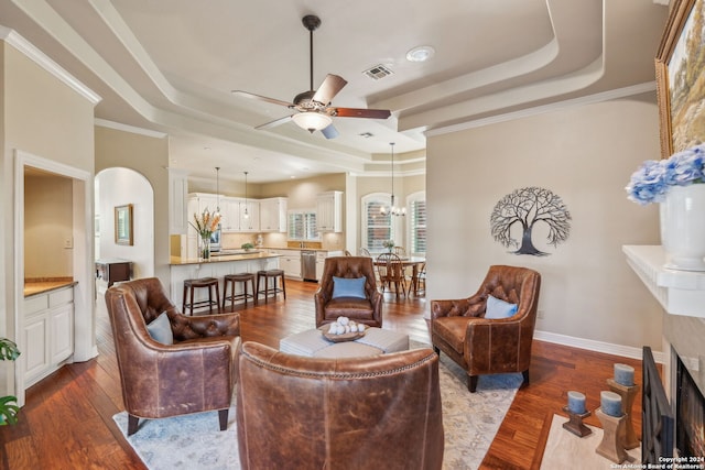 living room with crown molding, ceiling fan with notable chandelier, a raised ceiling, and light wood-type flooring