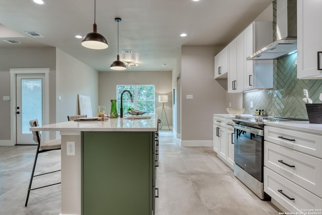 kitchen with stainless steel range with electric cooktop, hanging light fixtures, wall chimney exhaust hood, an island with sink, and white cabinetry