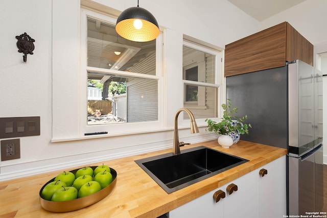 kitchen featuring sink, white cabinets, butcher block countertops, stainless steel refrigerator, and hanging light fixtures