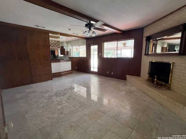 unfurnished living room with beamed ceiling, ceiling fan, a brick fireplace, and wood walls