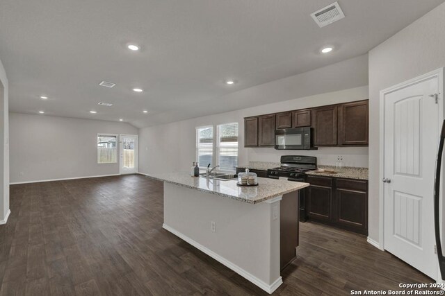 kitchen featuring dark wood-type flooring, light stone counters, black appliances, dark brown cabinets, and a center island with sink