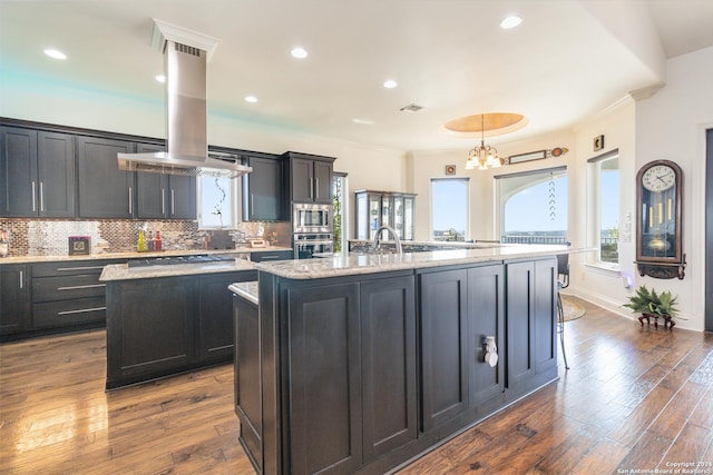 kitchen featuring a kitchen island with sink, ventilation hood, light stone countertops, appliances with stainless steel finishes, and dark hardwood / wood-style flooring