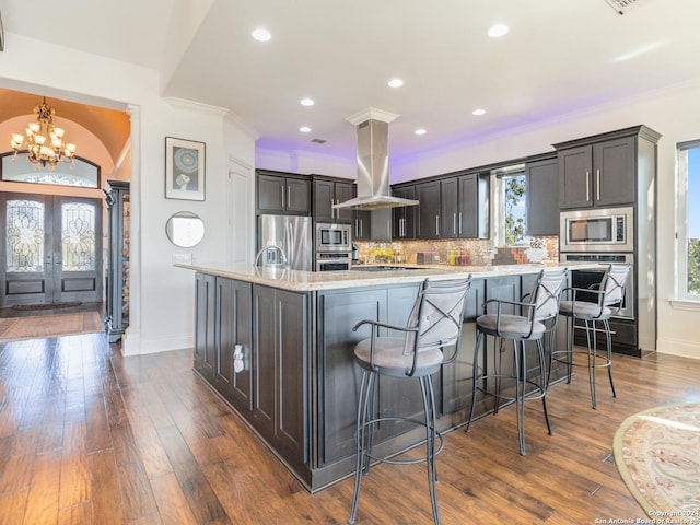 kitchen featuring island exhaust hood, appliances with stainless steel finishes, tasteful backsplash, a kitchen island with sink, and an inviting chandelier