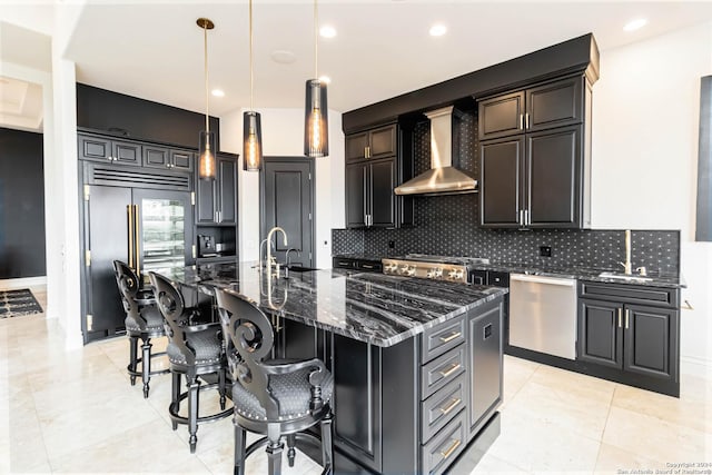 kitchen with an island with sink, appliances with stainless steel finishes, dark stone countertops, hanging light fixtures, and wall chimney range hood