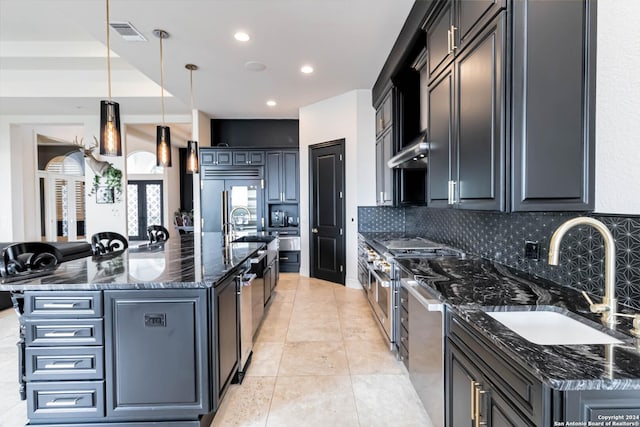 kitchen featuring a center island with sink, high quality appliances, dark stone countertops, and decorative light fixtures