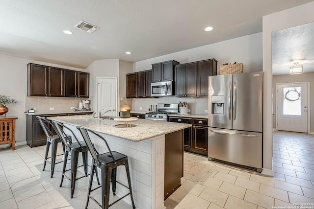 kitchen with a kitchen bar, decorative backsplash, light stone countertops, stainless steel appliances, and an island with sink