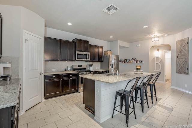 kitchen with dark brown cabinetry, light stone countertops, stainless steel appliances, and a kitchen island with sink