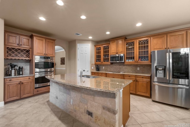 kitchen with appliances with stainless steel finishes, sink, backsplash, a kitchen island with sink, and light stone counters