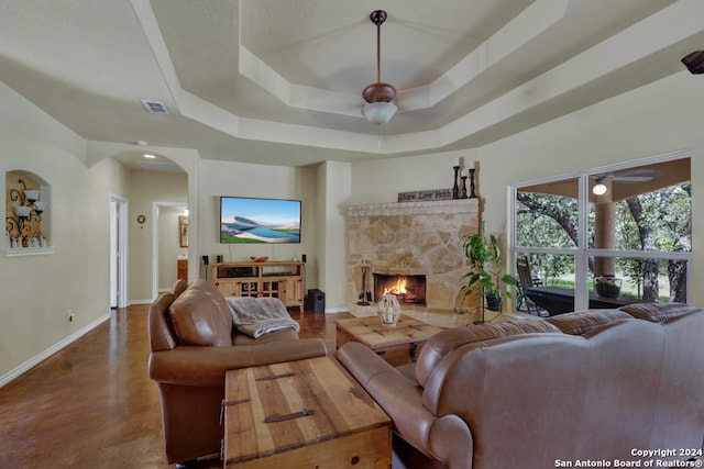 living room with a raised ceiling, a stone fireplace, and ceiling fan