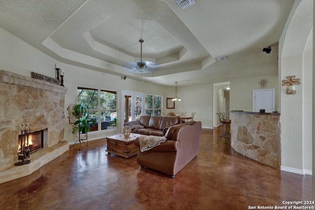 living room with a raised ceiling, a textured ceiling, a stone fireplace, and ceiling fan