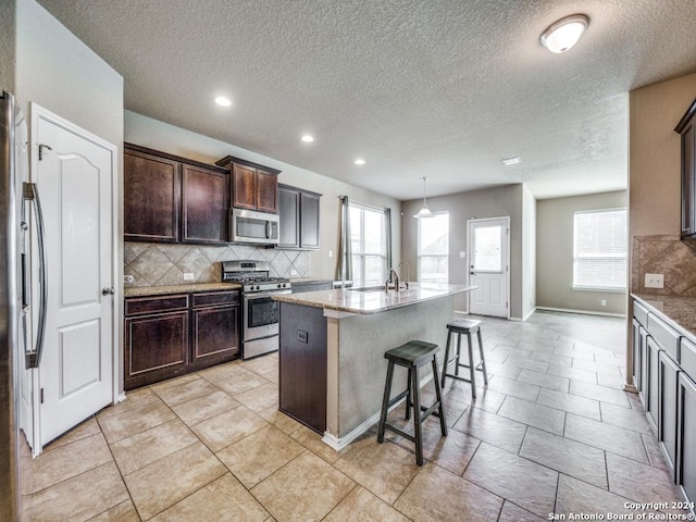 kitchen with appliances with stainless steel finishes, tasteful backsplash, dark brown cabinets, decorative light fixtures, and an island with sink