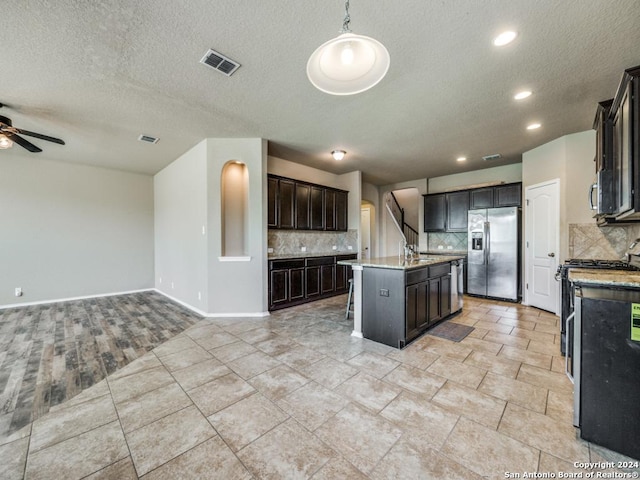 kitchen with pendant lighting, a center island with sink, a textured ceiling, appliances with stainless steel finishes, and tasteful backsplash