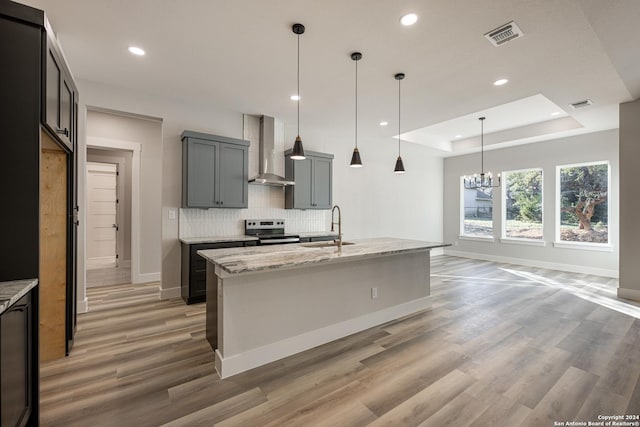 kitchen featuring wall chimney exhaust hood, light stone countertops, stainless steel range with electric cooktop, and an island with sink