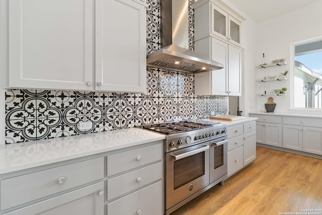 kitchen with backsplash, range with two ovens, wall chimney range hood, light hardwood / wood-style flooring, and white cabinetry