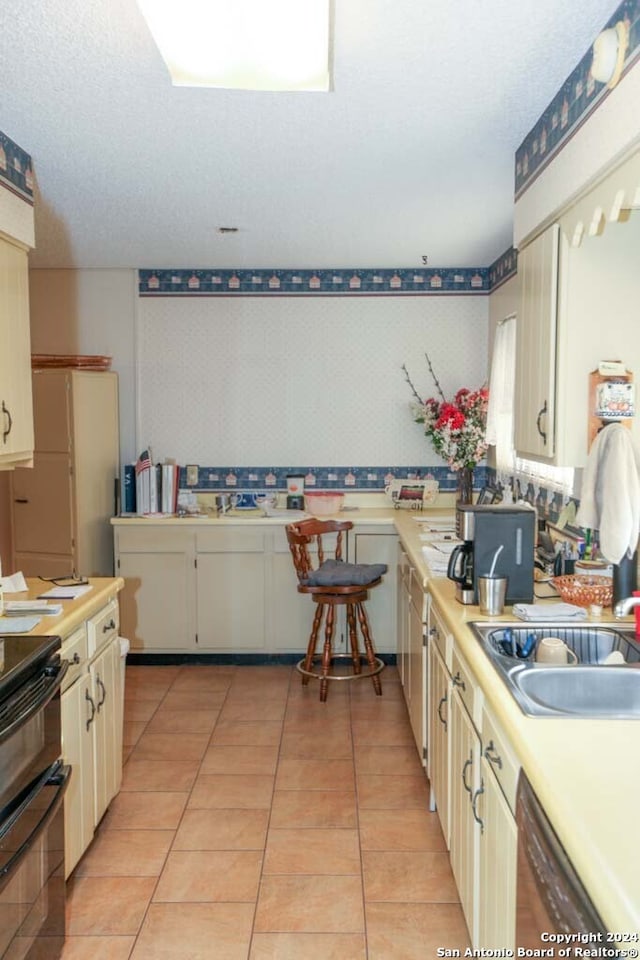 kitchen with dishwasher, black range oven, sink, light tile patterned floors, and cream cabinetry