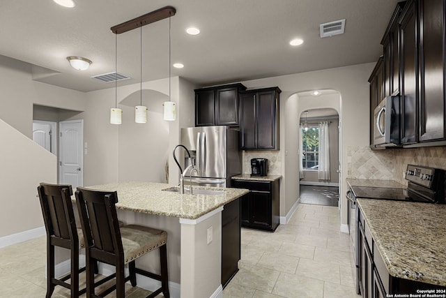 kitchen with light stone countertops, hanging light fixtures, tasteful backsplash, an island with sink, and appliances with stainless steel finishes