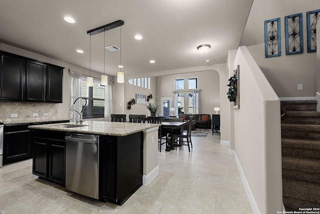 kitchen with dishwasher, light stone counters, backsplash, decorative light fixtures, and a center island with sink