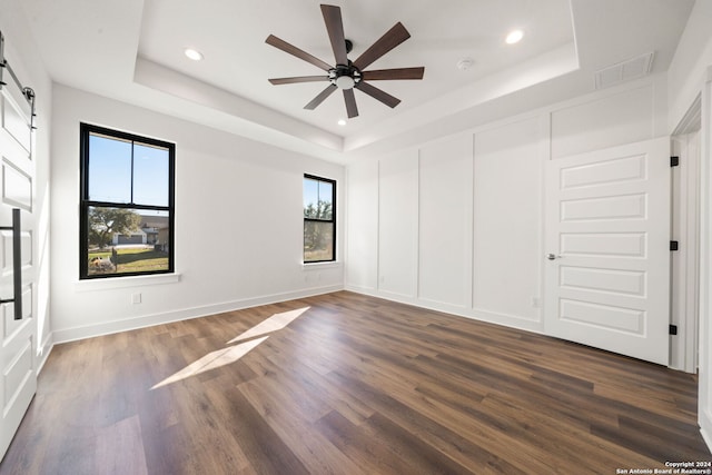 empty room featuring ceiling fan, a barn door, a raised ceiling, and dark hardwood / wood-style flooring