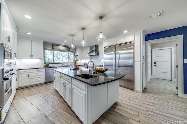 kitchen with built in appliances, wall chimney exhaust hood, white cabinets, and a kitchen island with sink