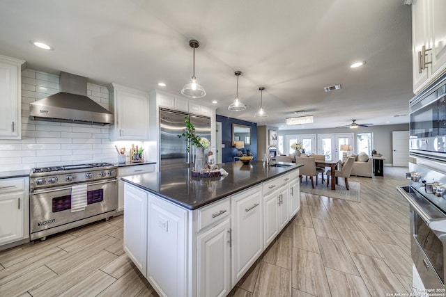 kitchen featuring wall chimney range hood, built in appliances, decorative light fixtures, white cabinetry, and an island with sink