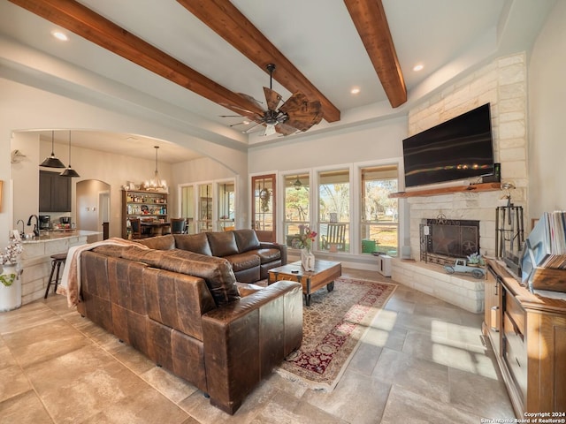 living room with a stone fireplace, beamed ceiling, and ceiling fan with notable chandelier