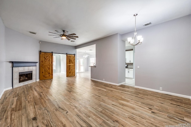 unfurnished living room featuring ceiling fan with notable chandelier, a barn door, light hardwood / wood-style floors, and a tiled fireplace