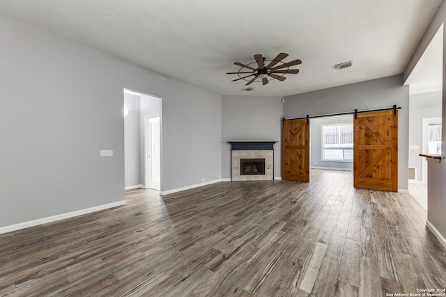 unfurnished living room featuring a barn door, ceiling fan, dark wood-type flooring, and a tiled fireplace