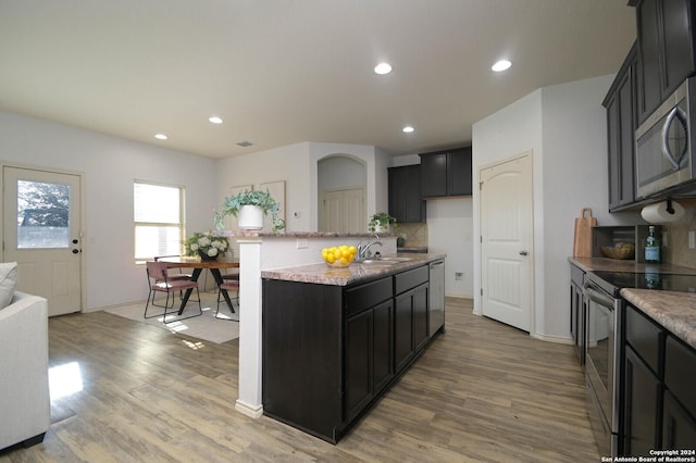 kitchen featuring sink, stainless steel appliances, light stone counters, wood-type flooring, and a center island with sink