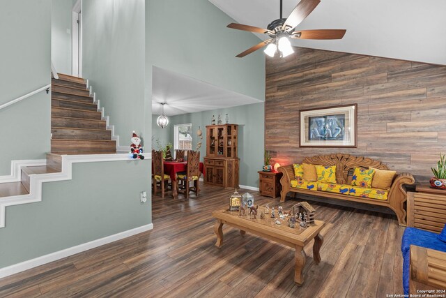 living room with high vaulted ceiling, ceiling fan with notable chandelier, dark wood-type flooring, and wooden walls