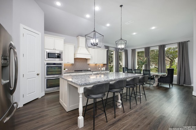 kitchen featuring pendant lighting, white cabinetry, appliances with stainless steel finishes, an island with sink, and custom range hood